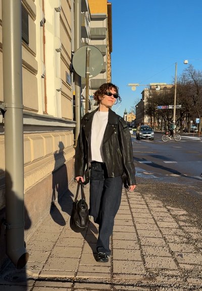 Chaqueta de motorista de cuero negro, camiseta blanca, jeans oscuros sueltos, llevando una bolsa negra. Cabello corto y rizado, gafas de sol. Entorno urbano.