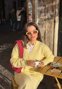 Cardigã amarelo claro em malha e calças combinando. Segurando uma chávena de café branca, sentado numa mesa amarela com produtos de maquilhagem próximos.