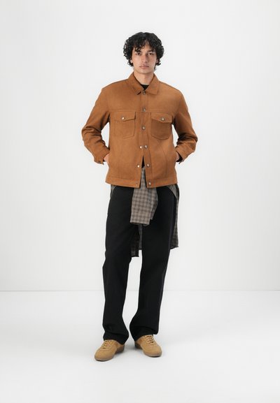 Chaqueta de ante marrón con dos bolsillos en el pecho, combinada con una camisa de cuadros gris y pantalones negros. El modelo lleva zapatillas beige. Diseño simple y casual.