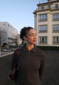 Le modèle porte une veste zippée à rayures foncées. Un petit sac bordeaux est suspendu à son épaule. En arrière-plan, des bâtiments modernes et des arbres.