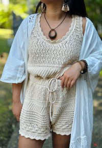 Γυναίκα με μπεζ ολόσωμη φόρμα χωρίς μανίκια πλεκτή, λευκό φουλάρι, μαύρο κρεμαστό κολιέ, σκουλαρίκια με φούντες και πολλά ξύλινα βραχιόλια, σε εξωτερικό χώρο.