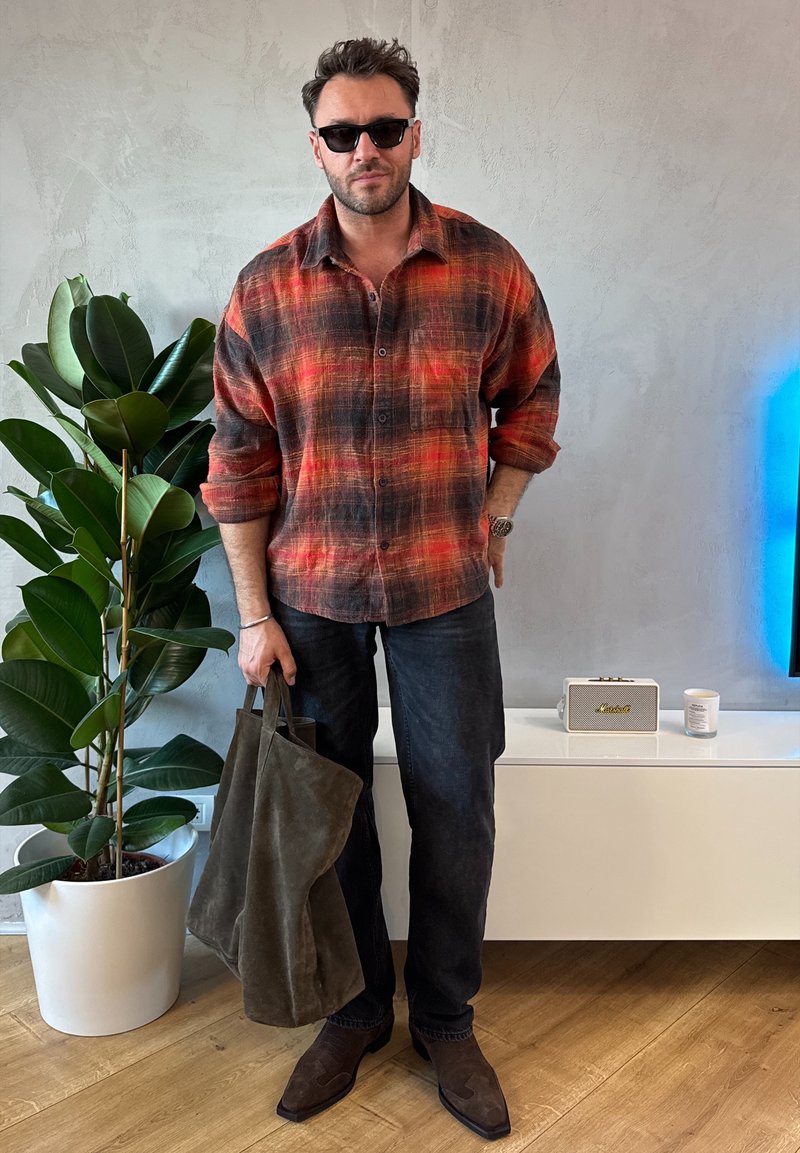 Kariertes rotschwarzes Flanellhemd, dunkle Jeans, olivgrüne Wildledertasche und braune Stiefel. Steht neben einer Pflanze und einem Lautsprecher in einem eleganten Interieur.