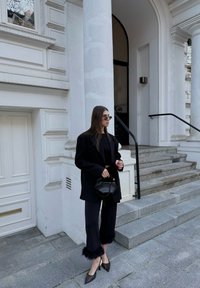 Μαύρο oversized σακάκι πάνω από ένα κοντό τοπ, σε συνδυασμό με παντελόνι wide-leg με φουντίτσες και στιλέτο τακούνια; κρατώντας μια μικρή μαύρη τσάντα.