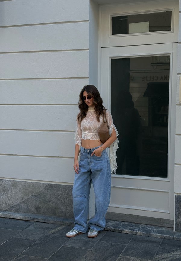 Femme aux cheveux ondulés portant des lunettes de soleil, un crop top en dentelle transparente, un jean large bleu et des baskets, posée contre un mur clair d’un bâtiment près d’une porte.