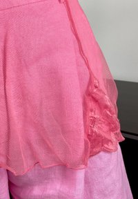 Zbliżenie różowej przezroczystej tkaniny z koronką kwiatową na ramieniu, na tle gładkiego różowego materiału teksturowego.