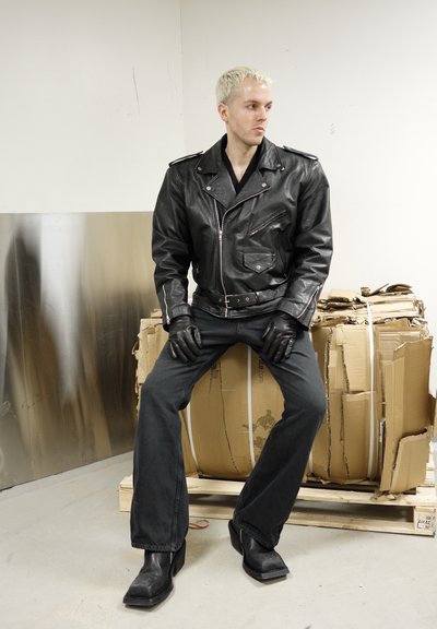 Persona con cabello rubio corto lleva chaqueta, guantes, pantalones y botas de cuero negro, sentada sobre cajas de cartón apiladas en una habitación blanca.