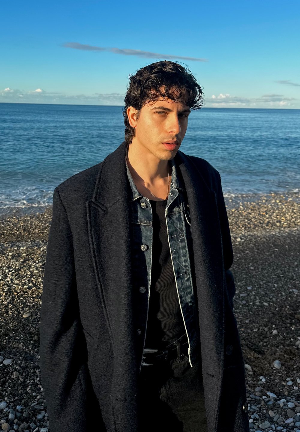 Homme portant un manteau en laine sombre par-dessus une veste en jean, une chemise noire et un pantalon, debout sur une plage rocheuse avec de l'eau en arrière-plan.
