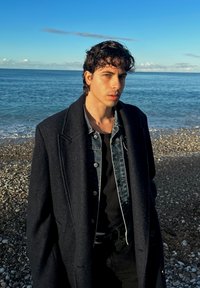 Homme portant un manteau en laine sombre par-dessus une veste en jean, une chemise noire et un pantalon, debout sur une plage rocheuse avec de l'eau en arrière-plan.