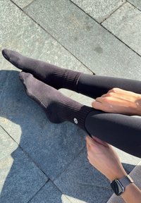 Fekete legényszemű zoknik bordázott textúrával, testhezálló kialakítással és logó részlettel, fekete leggingsszel viselve, texturált kőfelületen.