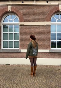 Groene leren jas met draakontwerp op de rug, bruine bontmuts, bruine rok, zwarte panty en bruine haklaarzen. Achtergrond van een bakstenen muur.