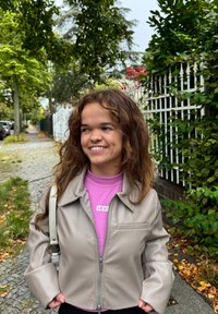 Beige imitationsläderjacka med dragkedja framtill, spetsig krage och en bröstficka, bärs över en rosa polotröja med en logotyp.