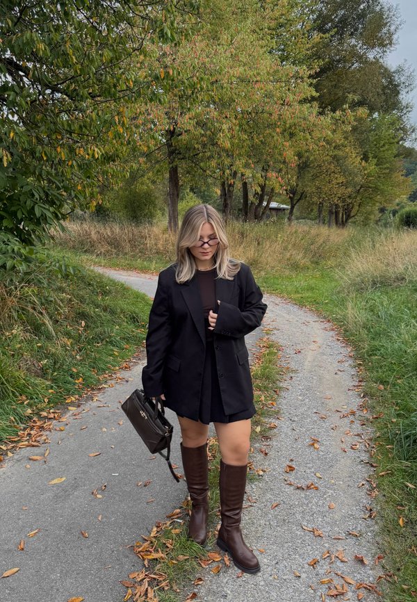 Mujer con blazer negro, falda y botas marrones caminando por un sendero cubierto de hojas rodeado de árboles verdes y otoñales.