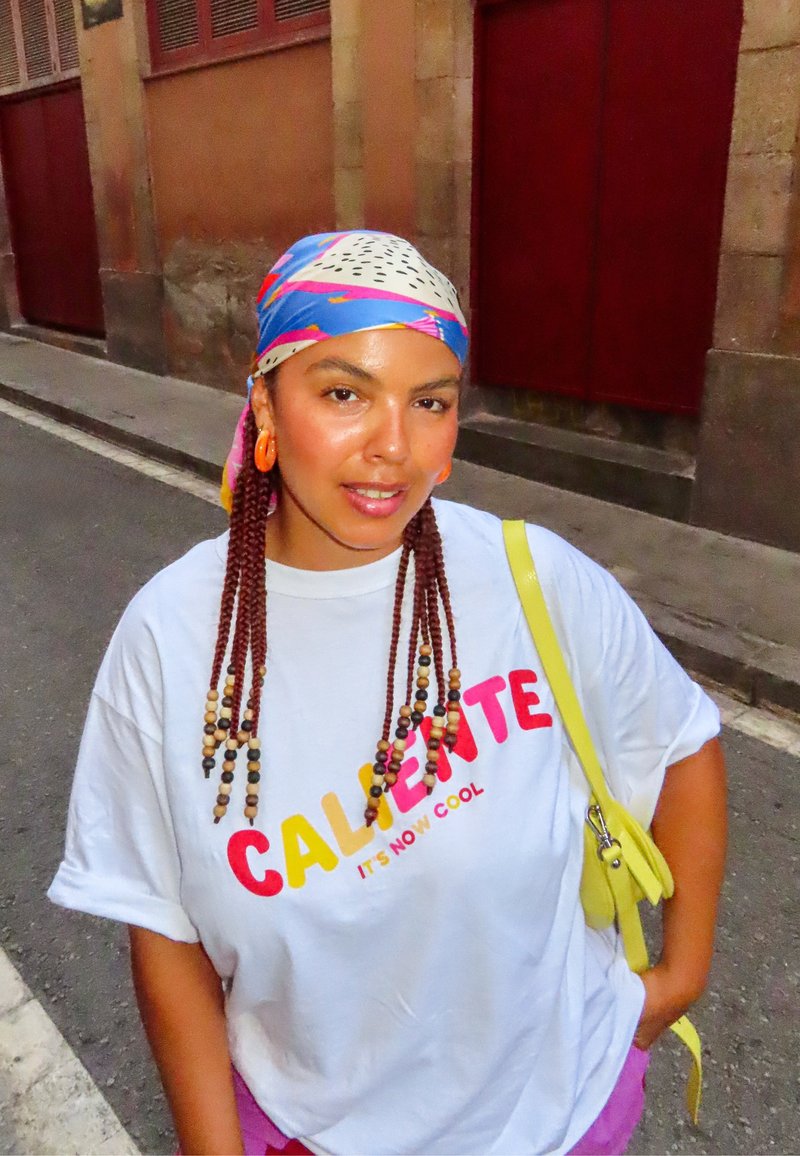 Donna con capelli intrecciati e perline colorate indossa una maglietta bianca "Caliente", fazzoletto decorato, orecchini arancioni e borsa gialla a tracolla, in piedi per strada.