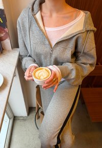 Personne en survêtement gris tenant une tasse de latte avec une mousse en forme de cœur, assise sur un tabouret en bois près d'une table en marbre.