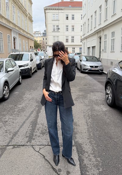 Blazer negro sobre una camisa blanca con cuello, combinado con jeans de pierna ancha azul y botas negras de punta, de pie en una calle de la ciudad.