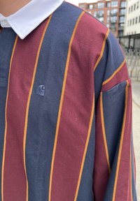 Polo a maniche lunghe con strisce verticali marrone, blu navy e oro, realizzato in cotone con colletto bianco e un piccolo dettaglio con logo.