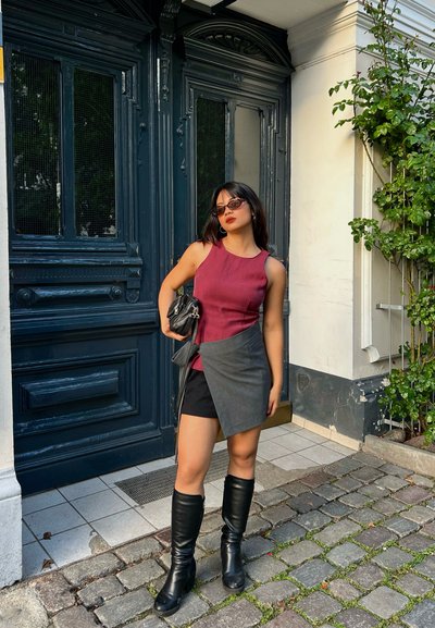 Top sin mangas color marrón sobre un falda envolvente gris, combinado con botas negras hasta la rodilla y un bolso negro. Fondo urbano con puerta decorativa.