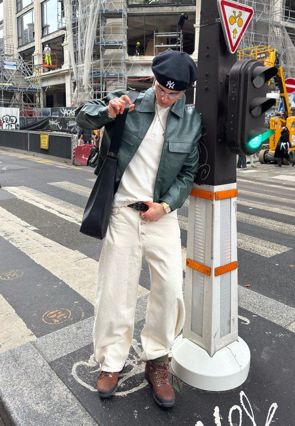 Oseba z rdečo baretko, zeleno usnjeno jakno, belimi hlačami in rjavimi škornji stoji na prehodu za pešce ob zeleni luči za pešce.