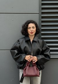 Donna con capelli ricci scuri che indossa una giacca di pelle nera e jeans chiari, che tiene una borsa bordeaux, in piedi contro un muro grigio.