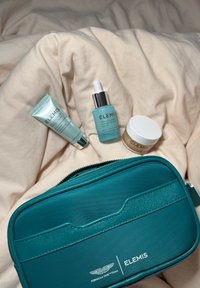 Sett med tre Elemis hudpleieprodukter: et lyseblått serum, en grønn krem og en sølvfarget tube, alle på en beige overflate. Inkluderer en teal glidelåspose.