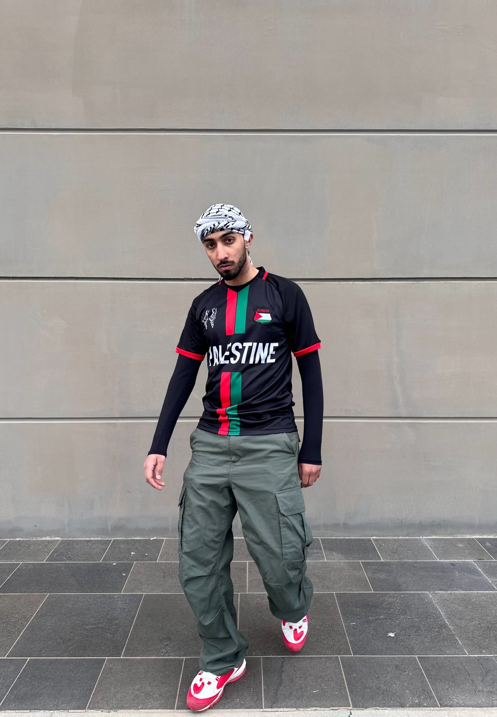Μαύρη ποδοσφαιρική φανέλα με κόκκινες λεπτομέρειες και πράσινη κατακόρυφη ρίγα, συνδυασμένη με φαρδιά πράσινα cargo παντελόνια και λευκά sneakers με κόκκινες λεπτομέρειες.
