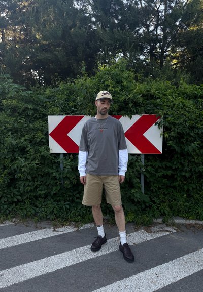 Un hombre que lleva una gorra, camisa gris sobre una camiseta blanca de manga larga, pantalones cortos beige y zapatos negros, está de pie en una carretera con señales rojas de flecha hacia la izquierda y densa vegetación detrás.