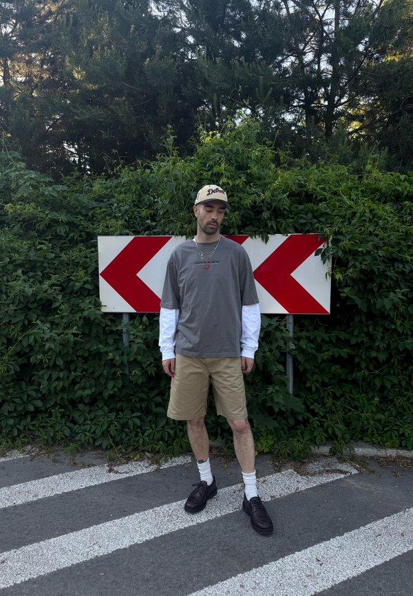 En mand iført en kasket, grå skjorte over en hvid langærmet, beige shorts og sorte sko står på en vej med røde venstre-pil skilte og tæt grøn vegetation bagved.