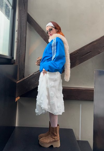 Chaqueta azul con rayas blancas, cuello de pelaje, falda blanca con flores, botines marrones, medias rosas con estampado, y gafas de sol.