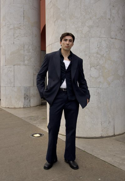 Traje navy con blazer de doble botonadura, pantalones a juego y botas negras. Camisa blanca visible debajo. Columnas de mármol en el fondo.