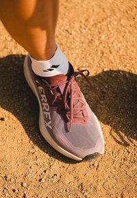 Lilla og grå sportssko med mesh-overdel, lisser og merket på siden. Brukt med en hvit sokk som har svarte detaljer på en sandaktig overflate.