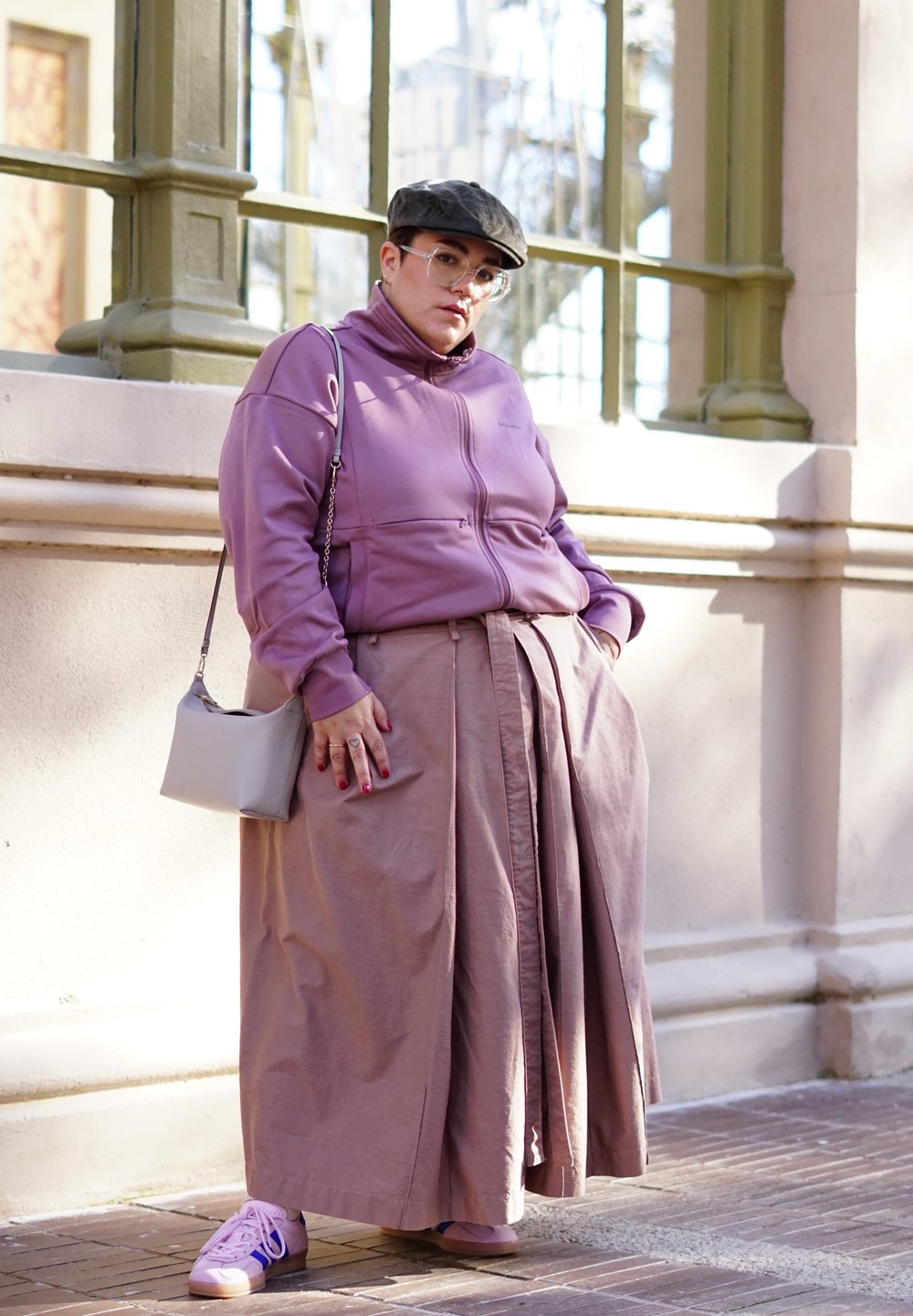 Giacca a zip viola chiaro, gonna lunga beige, sneaker rosa, borsa a tracolla grigia, occhiali e berretto piatto. Luce naturale, pavimentazione in mattoni.