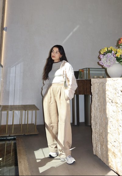 Pantalones de tiro ancho a rayas beige y un suéter ligero sobre una camiseta ajustada, de pie en un espacio minimalista con un arreglo floral y una mesa de cristal.