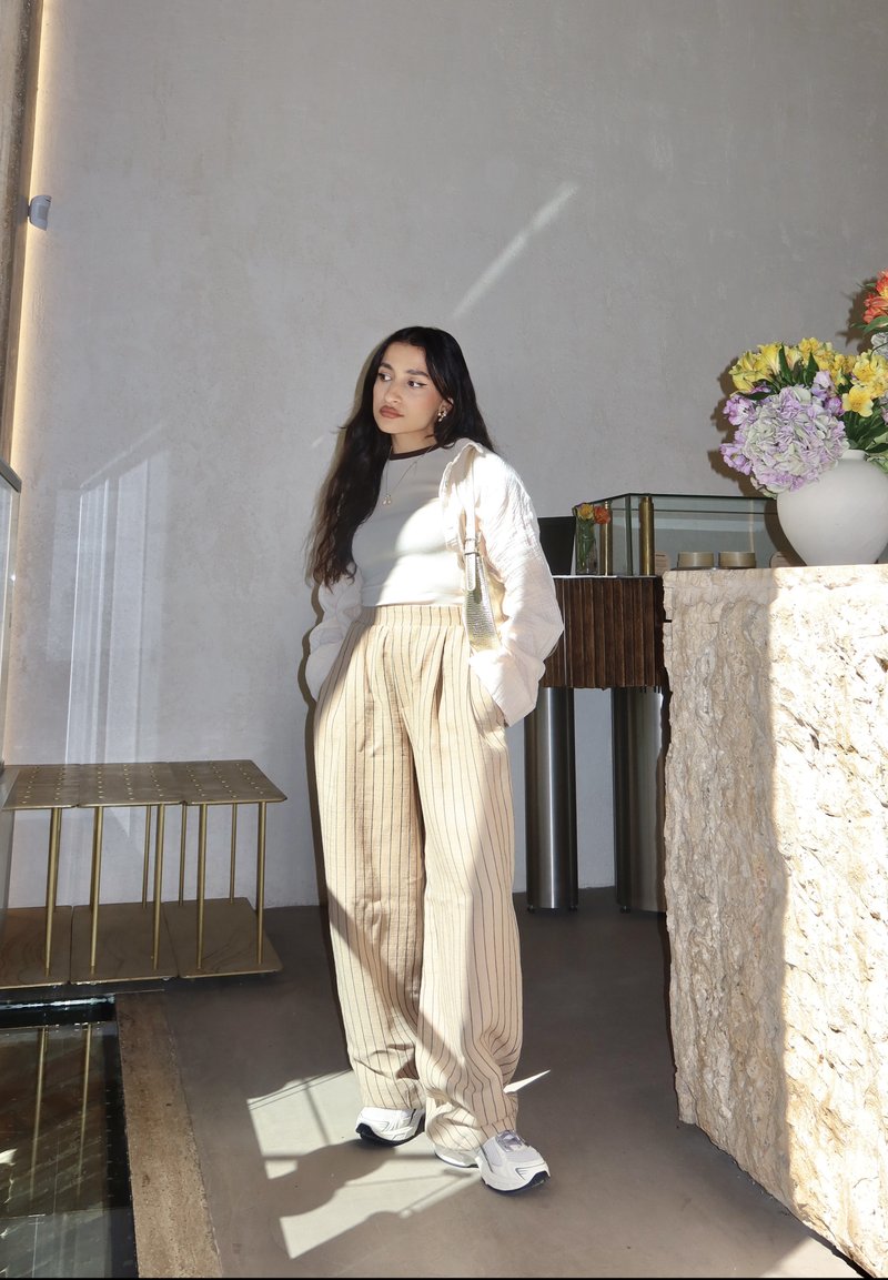 Beige striped wide-leg pants and a light sweater over a fitted top, standing in a minimal space with a floral arrangement and a glass table.