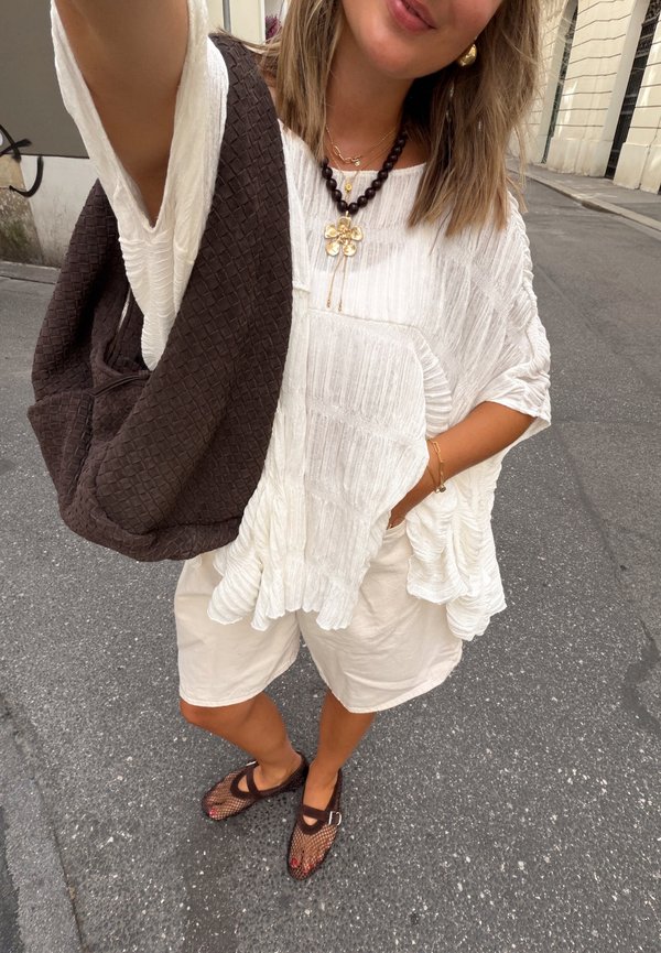 Frau in weißer, strukturierter Bluse und Shorts trägt eine dunkle Perlenkette mit Blumenanhänger, braune Maschenschuhe und trägt eine große braune gewebte Tasche.