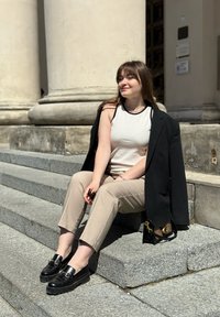 Musta bleiseri heitettynä hartioille, beige housut, valkoinen hihaton toppi ja massiiviset mustat loaferit kultaisilla yksityiskohdilla, istumassa kivisillä portailla.