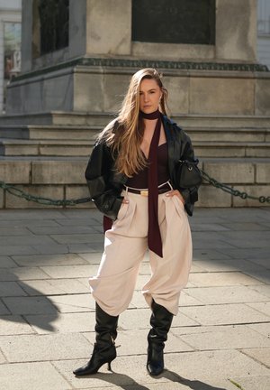 Mujer de cabello largo con chaqueta de cuero negra, blusa burdeos y bufanda, pantalones anchos beige y botas negras de tacón, de pie sobre un pavimento de piedra.