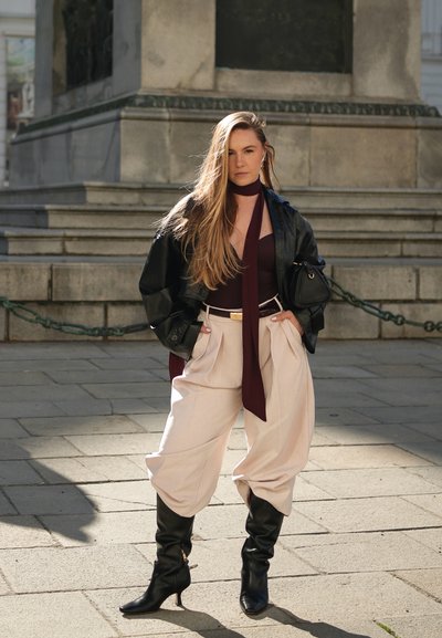 Mujer de cabello largo con chaqueta de cuero negra, blusa burdeos y bufanda, pantalones anchos beige y botas negras de tacón, de pie sobre un pavimento de piedra.