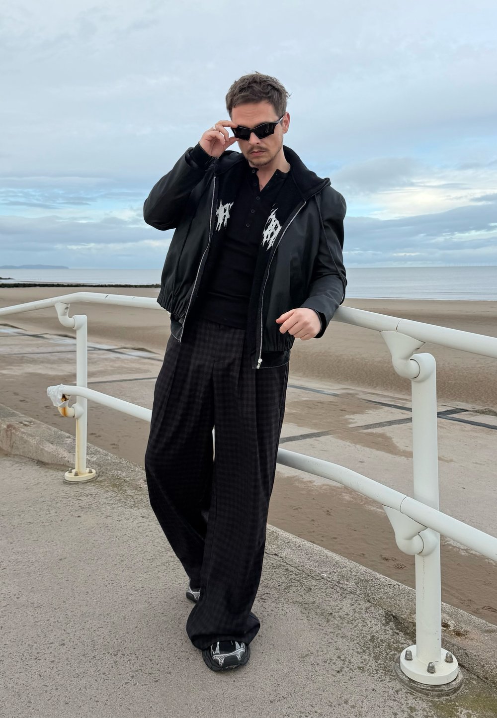 Crna kožna bomber jakna preko crne polo majice s bijelim detaljima, uparena s širokim crnim hlačama na kockice i sportskim crnim cipelama.