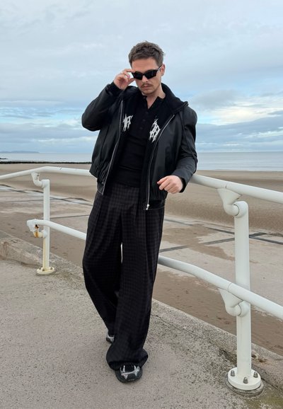 Cazadora bomber de cuero negro sobre un polo negro con detalles blancos, combinada con pantalones anchos negros a cuadros y zapatos deportivos negros.