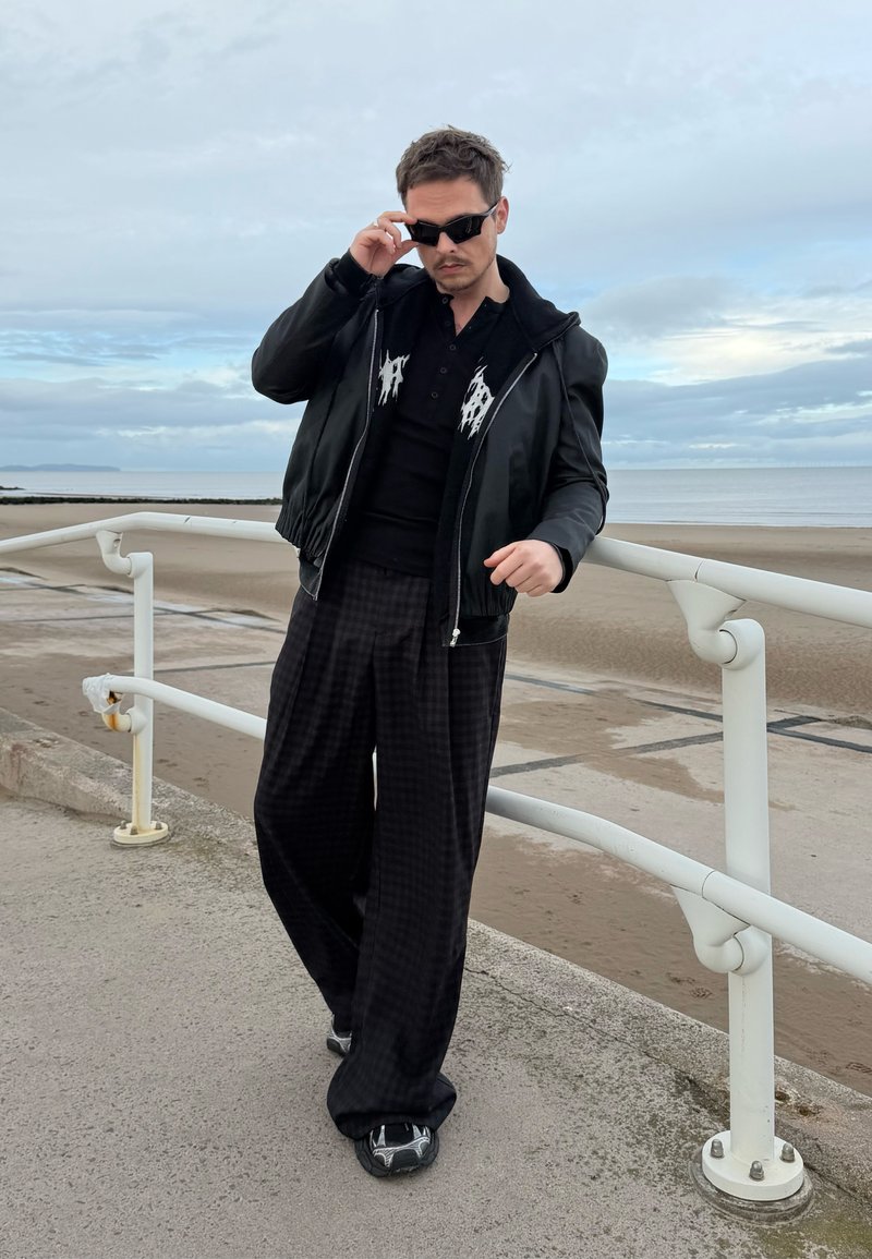 Geacă bomber neagră din piele peste un polo negru cu accente albe, asortată cu pantaloni largi negri în carouri și pantofi sport negri.