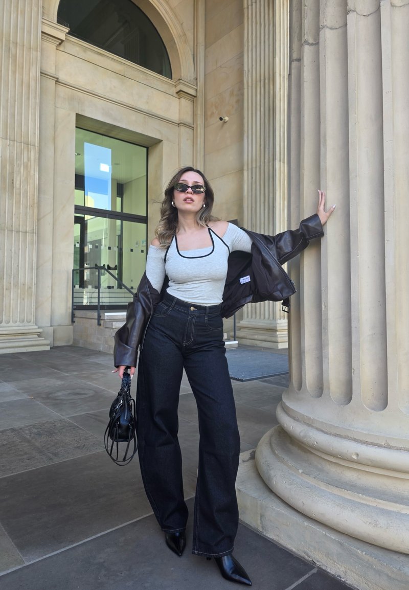 Haut gris à épaules dénudées avec bord noir, associé à un jean foncé taille haute. Superposé avec une veste en cuir marron, tenant un sac à main noir.