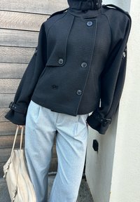 Sort dobbeltradet jakke med tekstureret finish, brede ærmer og store knapper, kombineret med lysegrå plisserede bukser og en beige taske.