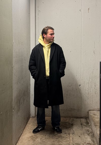 Un abrigo de lana negro sobre una sudadera amarilla, combinado con jeans oscuros y zapatillas negras. El entorno cuenta con paredes y suelo de hormigón.