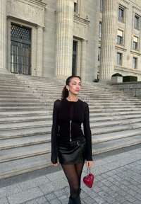 Črn dolg rokav majica z zadrgo, v kombinaciji s črnim usnja mini krilom in rdečo torbico v obliki srca. Kamnite stopnice in klasična arhitektura v ozadju.