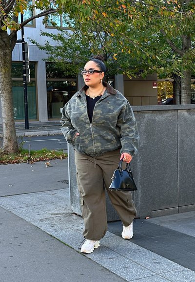 Chaqueta bomber de camuflaje con cuello marrón, parte superior negra y pantalones cargo verdes. Bolso negro, zapatillas blancas, de pie en una acera.