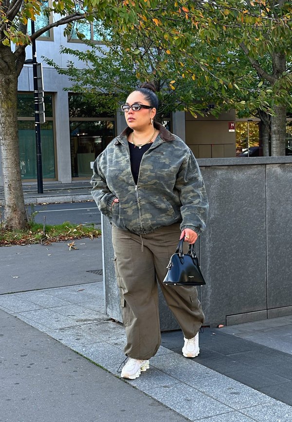 Casaco bomber camuflado com colarinho castanho, parte superior preta e calças cargo verdes. Bolsa preta, ténis brancos, em pé numa calçada.