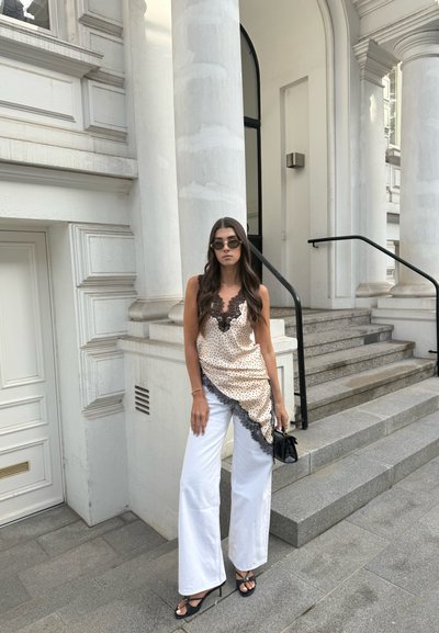 Mujer con gafas de sol, blusa beige con lunares y detalles de encaje, pantalones blancos de pierna ancha y tacones negros, de pie junto a escalones y columnas de piedra.