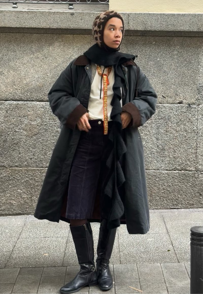 Femme portant un manteau long, des bottes noires, un foulard léopard et une écharpe noire, debout contre un mur en pierre sur un trottoir.