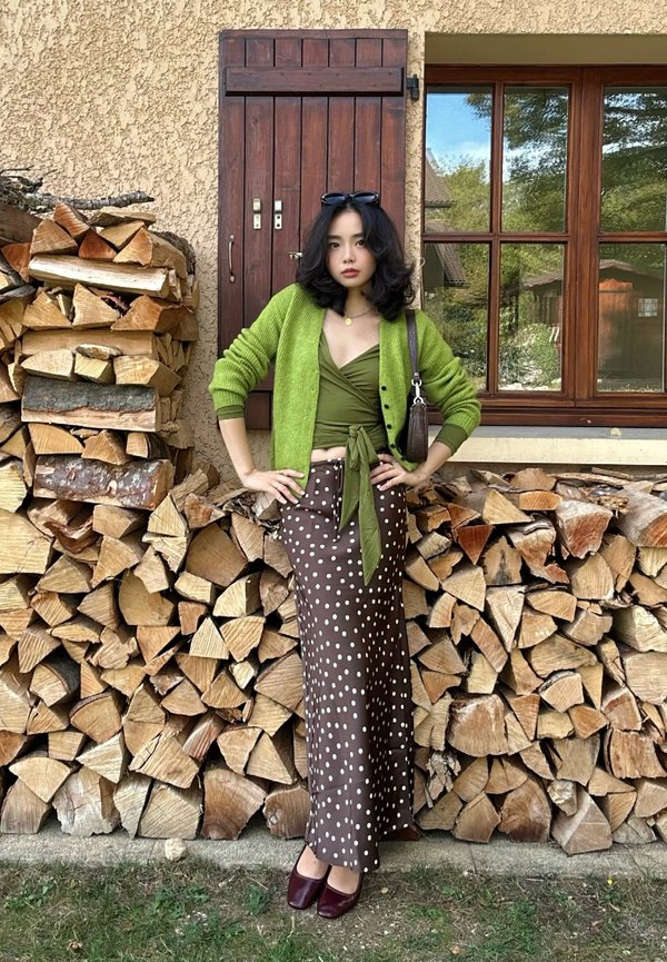 Jeune femme en cardigan vert et jupe à pois se tient les mains sur les hanches devant du bois de chauffage empilé et une fenêtre rustique.