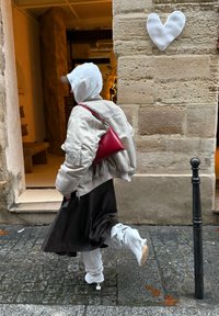 Personne portant un sweat à capuche blanc, une veste bomber beige, une jupe marron et des bottes blanches, portant un sac rouge, marchant sur une rue pavée à côté d'un mur en pierre avec un cœur.